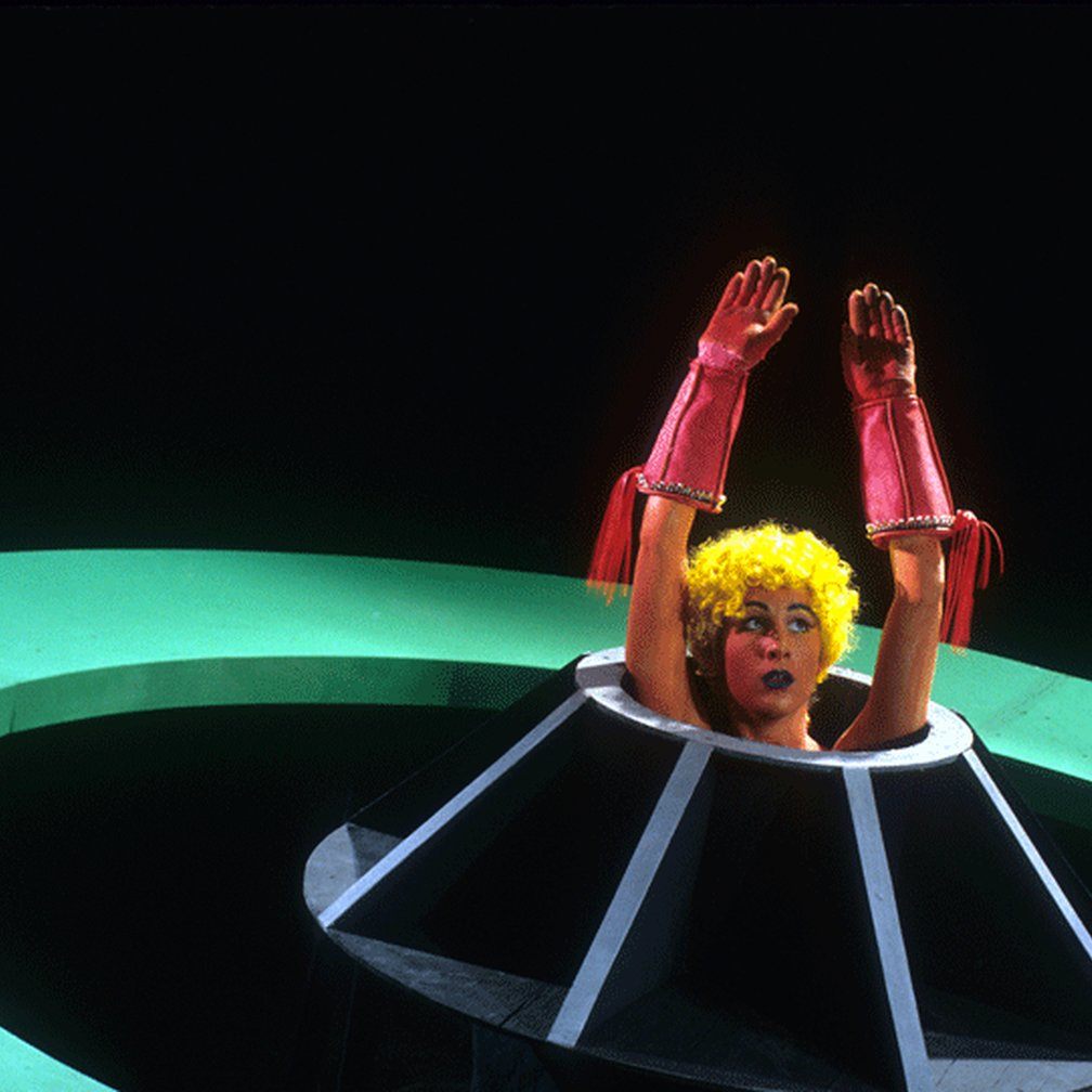 Woman with pink gloves and a yellow wig emerging from spaceship